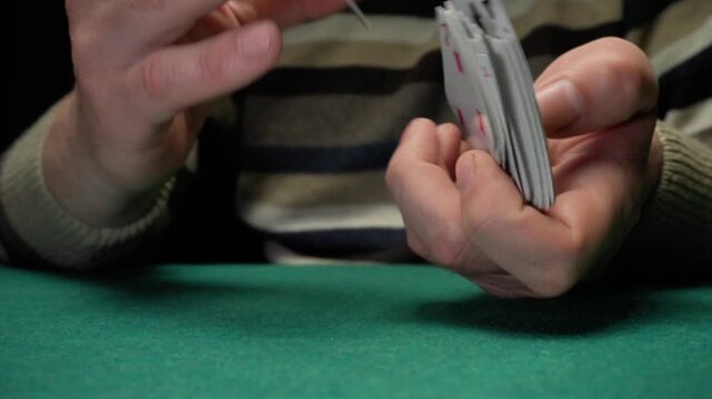 Hands of a Caucasian man shuffle playing cards on a green felt table, showcasing a focused card manipulation technique in a dimly lit setting