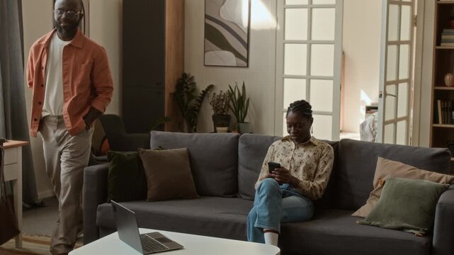 Medium full shot of cheerful African American man walking into living room, playing guess game with wife sitting on couch, presenting with surprise holiday trip tickets, woman cheering and hugging him