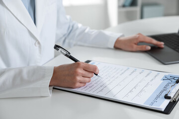 Fototapeta premium Doctor in medical coat working with clipboard and laptop at white table in clinic, closeup