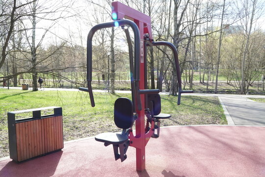 Professional red and black outdoor chest press fitness machine on a dedicated sports ground in a public park area with green grass and trees in the background under bright spring sunlight flare.