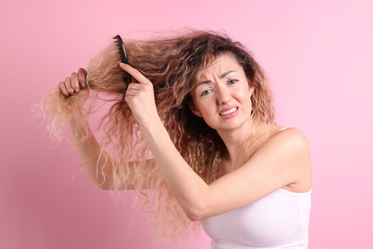 Unhappy woman trying to brush her tangled hair on pink background