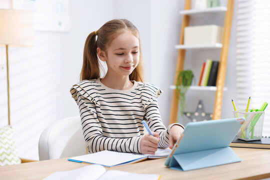 Cute girl with tablet doing homework at table indoors