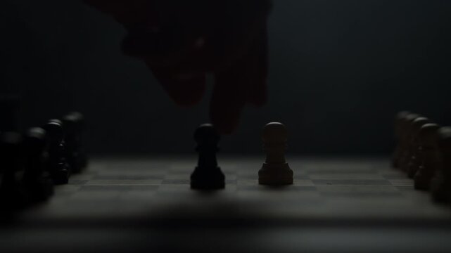 Cinematic close-up of a hand moving a chess piece in a dark atmospheric room. Low-key lighting highlights strategy, planning, and intellectual competition. Dramatic silhouette of a chess game.