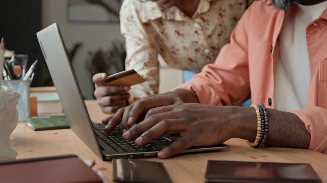 Close-up of hands of unrecognizable black couple purchasing vacation tickets online with credit card, man typing on laptop computer while woman reading aloud number
