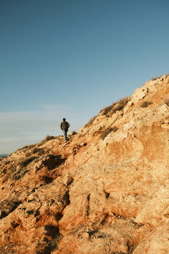 Lone hiker ascends a rugged, sun-drenched rocky hillside under a clear blue sky