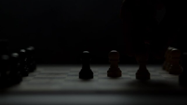 Cinematic close-up of a hand moving a chess piece in a dark atmospheric room. Low-key lighting highlights strategy, planning, and intellectual competition. Dramatic silhouette of a chess game.