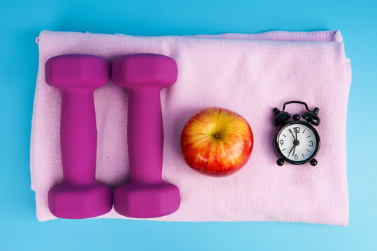 Healthy lifestyle and fitness routine concept with dumbbells, apple, and alarm clock on towel, symbolizing exercise, diet, time management, weight loss, and overall wellness.