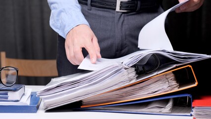 Business person sorting through a large stack of documents and binders on a desk, representing administration, paperwork, workload, and office organization