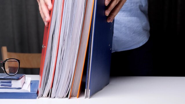 Person arranging a large stack of heavily filled binders and folders on a white desk, representing business organization, administration, and bureaucracy