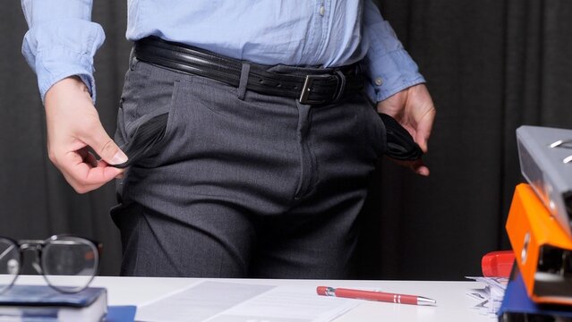 Businessman standing over a cluttered desk, pulling out both empty pockets from his formal trousers, symbolizing financial distress, poverty, and debt