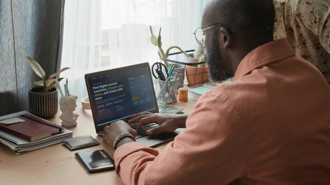 Medium rear shot of young African American husband and wife choosing travel dates on flight booking website on laptop, while preparing to purchase tickets for summer holidays overseas