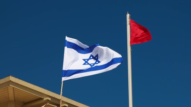 Israeli national flag waving beside a red warning flag against a clear blue sky. A scene above a beach structure roof indicating safety status or caution in the coastal area of Israel, sunny day