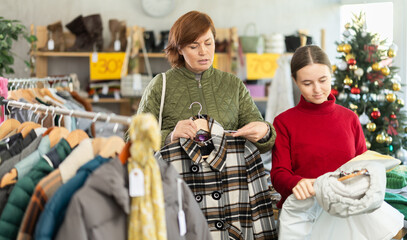 Fototapeta premium In sales area of store during Christmas sale, woman with teen daughter customer choose coat or jacket. Creating capsule wardrobe, branded clothing at affordable prices. Modern urban fashion