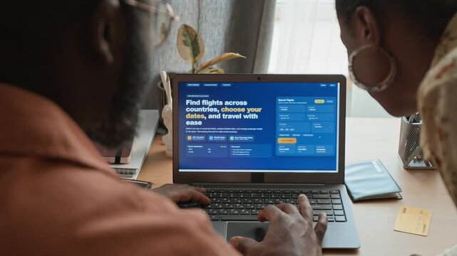 Rear over-shoulder shot of young black couple choosing travel dates and destination on flight reservation website on laptop, then deciding to upgrade service class for luxury experience