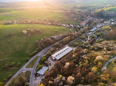 Aerial view of a large agricultural building and winding roads surrounded by blossoming trees and green rolling hills during golden hour in Cerin, Banska Bystrica Region, Slovakia.