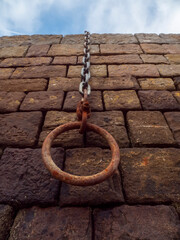 Fototapeta premium Rusty chain hangs from a weathered stone wall or a pier in a port. Each brick bears age, moss, and sun-worn texture. Chain links sag gently, offering a sense of decay.