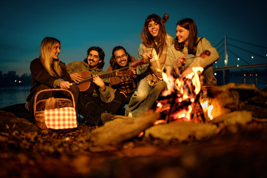 Friends gather around a campfire by a river while playing music and enjoying a picnic during the evening