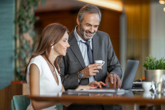 Business meeting occurs in an office with two people discussing work while using a laptop and enjoying coffee in the morning