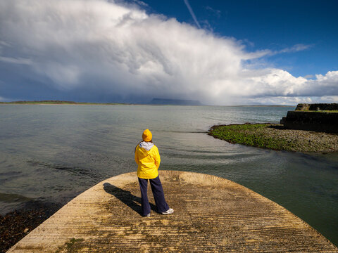 Lone figure stands on a weathered pier, gazing at the sea. Bold clouds loom overhead, casting dramatic shadows. Model in yellow travel jacket. Sligo, Ireland. Benbulbin mountain in the background.