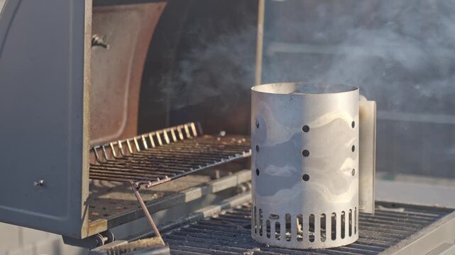 Smoking charcoal chimney starter stands on barbecue grill outdoors. Concept of grilling preparation and barbecue setup