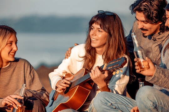 Group of friends play music and enjoy drinks by the water during golden hour in a casual outdoor setting