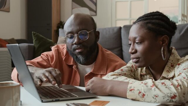 Medium close-up of African American husband and wife searching for travel options on laptop, choosing convenient flights for low price on booking aggregator website, wallet and credit cards on table