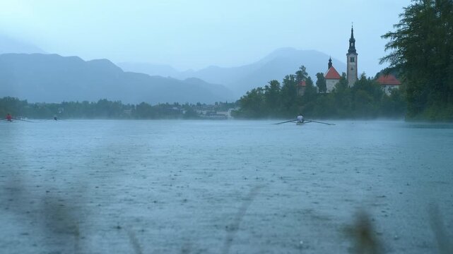 Rowers training on lake bled in the rain with island church in background. Rowing athletes training on lake bled during rainy weather, revealing picturesque slovenian landscape with island church