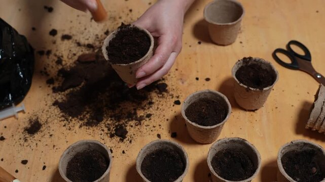 Girl gardener fill a peat pot with dark soil 4k video. Gardener scoops rich earth into a biodegradable cup using a wooden-handled shovel. Filling Seedling Pots with Soil.