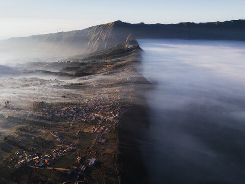 Aerial view of Bromo Tengger Semeru National Park with the village of Cemoro Lawang perched on a cliff edge above a thick sea of clouds at sunrise Bromo, East Java, Indonesia.