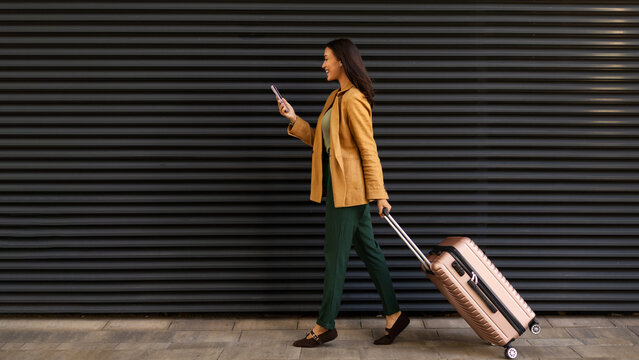 Young caucasian female traveler walking with rolling suitcase while looking at phone