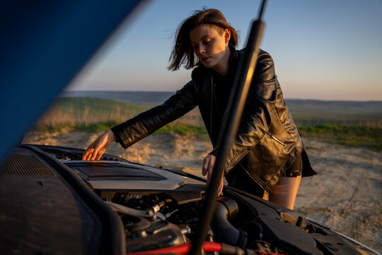 Woman inspecting engine compartment under open car hood outdoors, checking vehicle condition in warm evening sunlight during roadside stop.