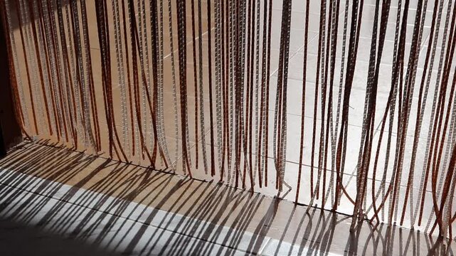 Plastic curtain strands sway in the breeze at a doorway, casting moving shadows on a tiled floor. Sunlit terrace visible beyond creates a calm, minimal summer scene.