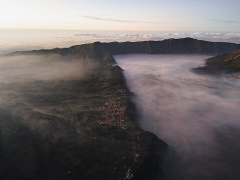 Aerial view of Bromo Tengger Semeru National Park with thick morning mist filling the volcanic caldera, rugged mountain ridges, and a small village at dawn Bromo, East Java, Indonesia.