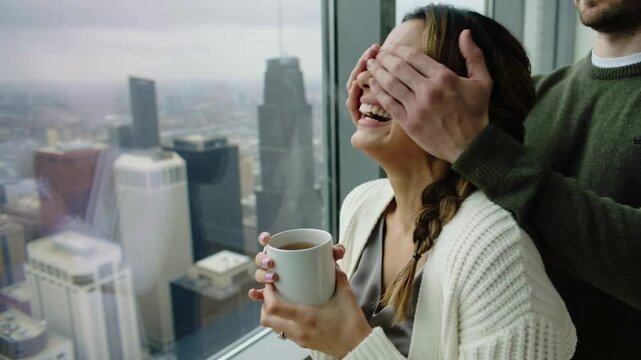 Young woman drinking tea while man covers her eyes in high rise apartment with city view, medium shot