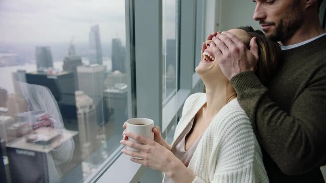 Man covering eyes of woman holding cup by window in high-rise apartment with city view, medium shot
