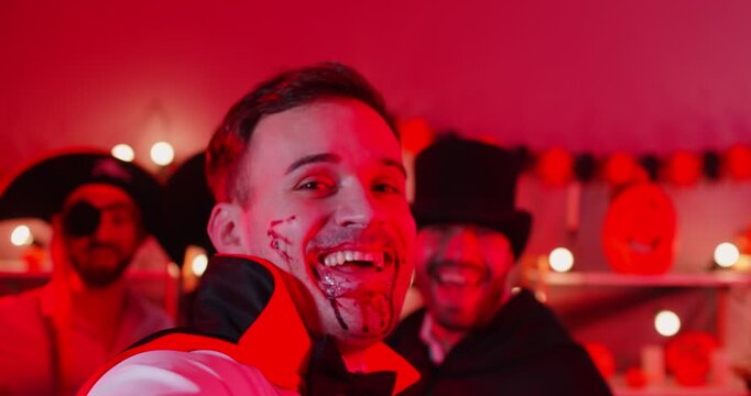 Cheerful man with bloody scars on face and his friends posing together for making selfie while being on party and celebrating Halloween, wearing spooky costumes, look happily at camera