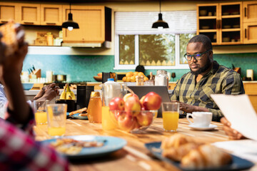 Fototapeta premium Black working dad on computer ignoring son and family at breakfast table, absorbed disconnected man distracted by his remote work. Inattentive parent distant with his children and wife.
