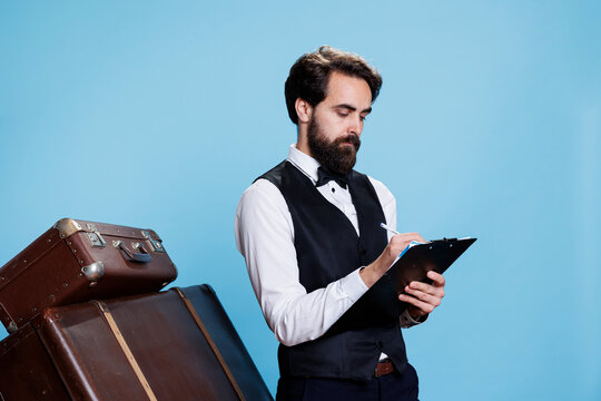 Hotel porter write information on files, filling in record papers with guest names for inventory. Professional bellboy checking list on clipboard documents, stand next to luggage.