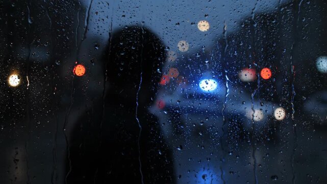 Man standing by window looking at rainy city street. Silhouette of head observing bokeh traffic lights. Cinematic storm mood in dark evening urban environment.