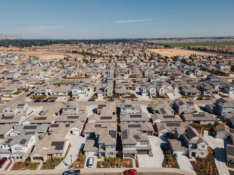 Aerial Planned Grid Community Long Perspective Revealing Symmetric Rows Of Houses, Narrow Backyards, Parked Cars, Distant Farmland, Serene Afternoon Light And Efficient Street Layout.