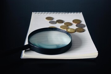A magnifying glass resting on a spiral notebook with various Malaysian coins, representing financial analysis, audit, and savings concepts. © Akram