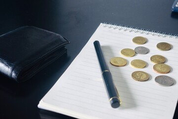 Side perspective of a closed black leather wallet next to a notebook with scattered coins and a pen on a dark surface. © Akram