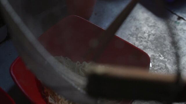 Boiled instant noodles drained in a red bowl, steamy Asian street food preparation close-up with strainer and chopsticks.