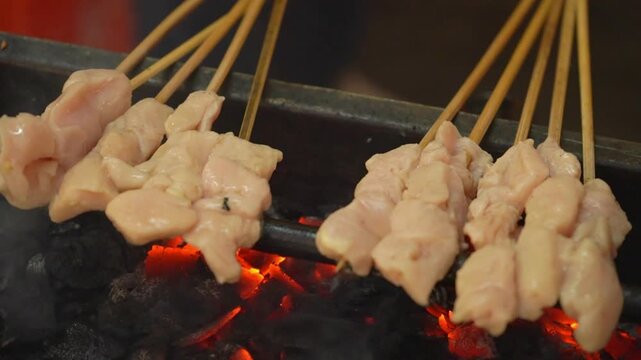 Chicken satay skewers grilling over smoky charcoal, raw marinated meat cooking on open fire in an Asian street food BBQ scene.