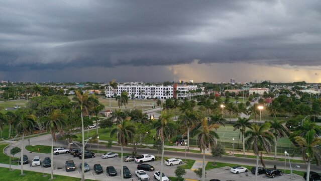 Aerial view of a massive shelf cloud and dark storm front looming over a suburban neighborhood featuring palm trees, a large parking lot, and a modern white apartment building.