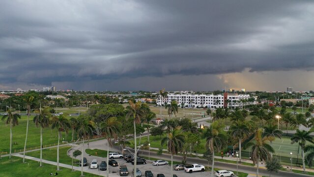 Aerial view of a suburban park with palm trees and a parking lot under a dramatic storm front with dark clouds and rain in the distance.
