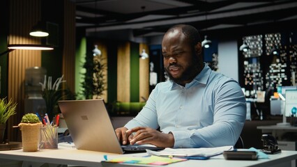 Fototapeta premium African american man does critical thinking and proofreading with papers before transcribing data on notebook, sitting in the office at night. Worker documenting important ideas.