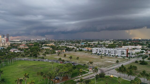 Aerial view of a massive shelf cloud formation approaching a suburban neighborhood with lush green parks, palm trees, and residential buildings under a dark stormy sky.