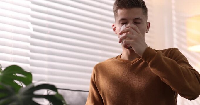 Man with glass of water taking pill near window indoors