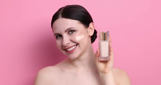 Beautiful woman with bottle of foundation and smear on her face against pink background